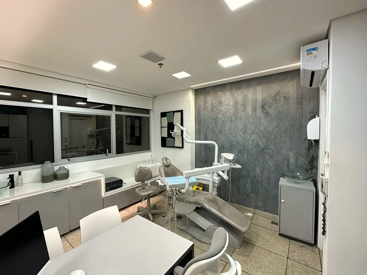 Sala de atendimento: cadeira odontológica, parede cinza em espinha de peixe, bancada clara e a mesa de conversa